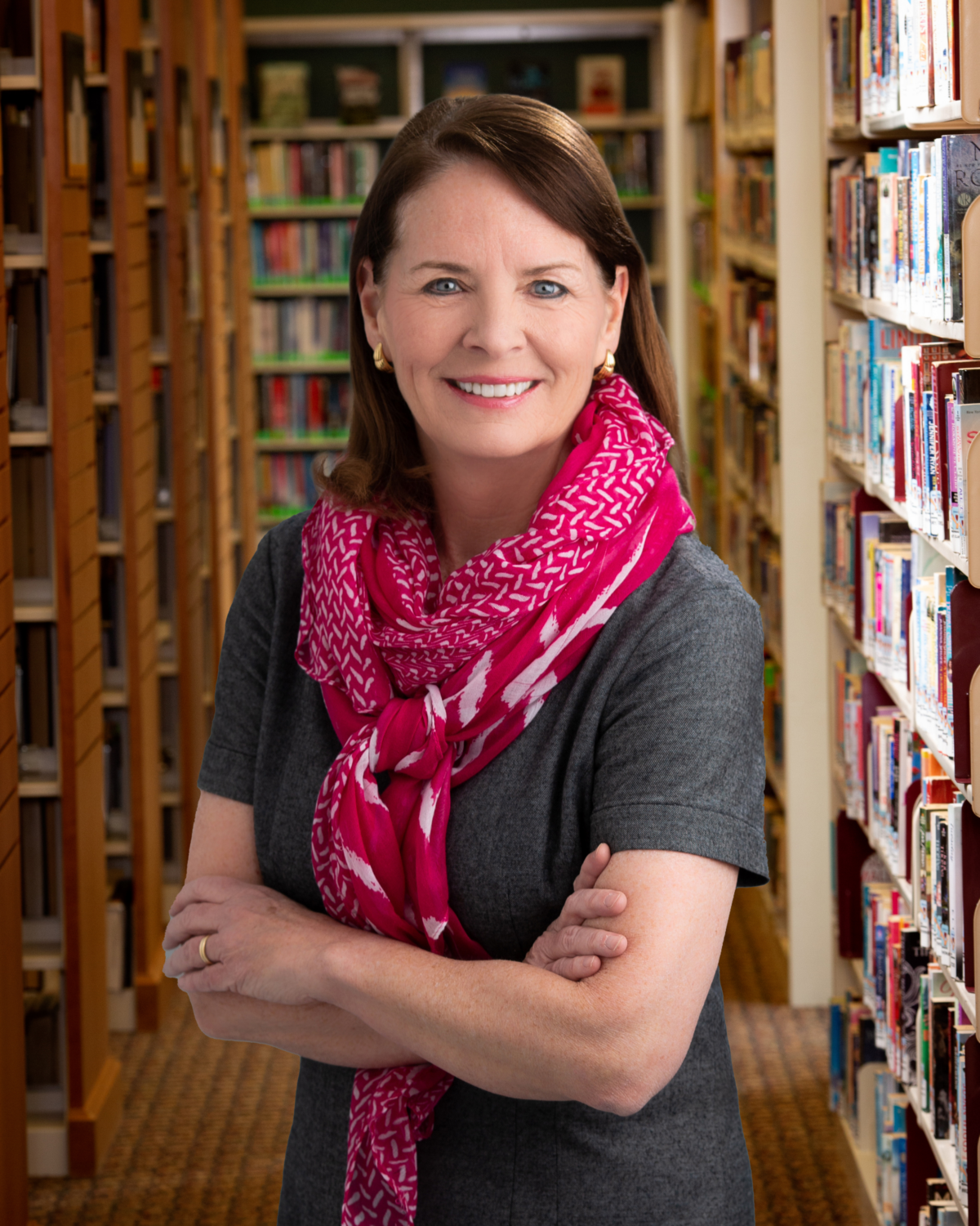 woman with pink scarf crosses her arms while standing in front of book shelves