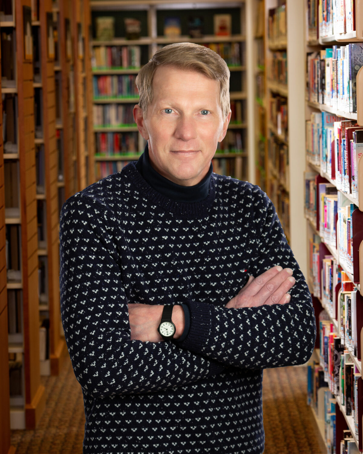 man with blue sweater crosses arms while standing in front of library shelves