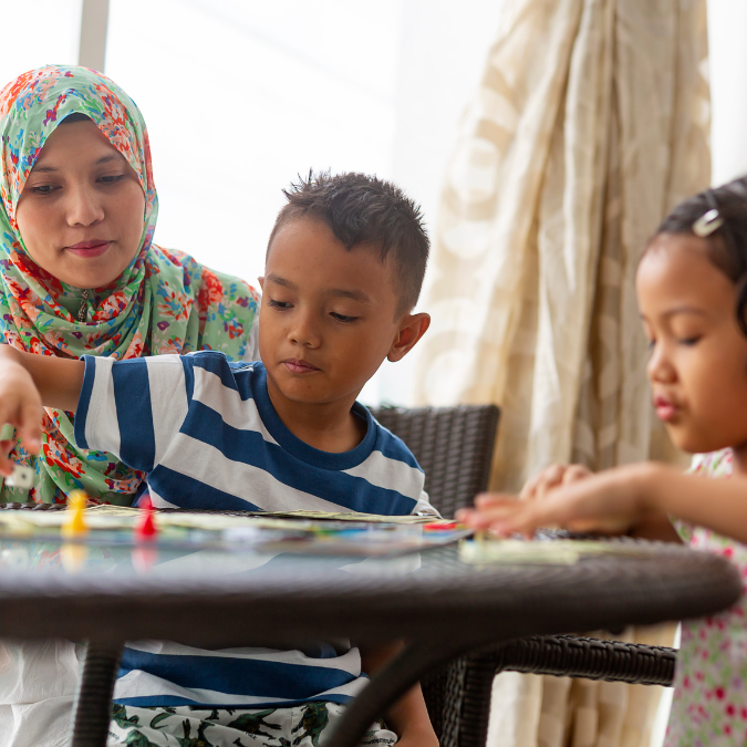 a women with a hijab plays board games at a table with a young boy and girl