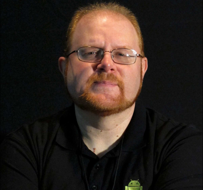 Image of Michael McEvoy, participant in Human Library and member of the Northville Public Library in Michigan. Man wearing glasses, crew cut red hair and beard with black shirt and dark background. Small smile. 