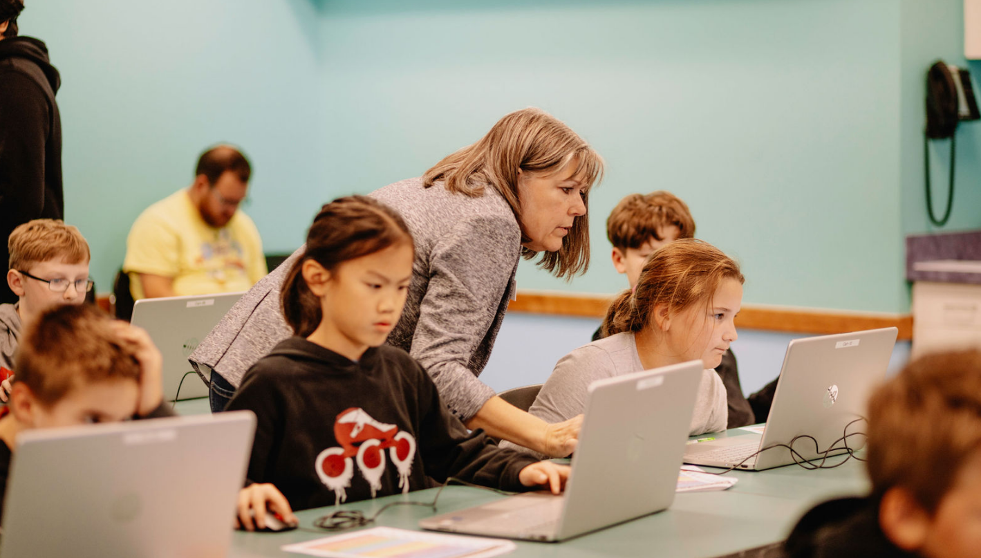 teacher leaning over a computer to help kids during a computer class
