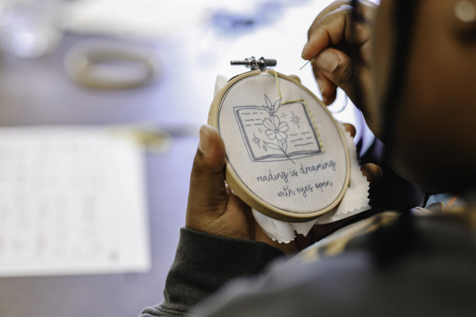 A patron embroiders fabric with the saying "Reading is dreaming with eyes open" with an open book above the text.