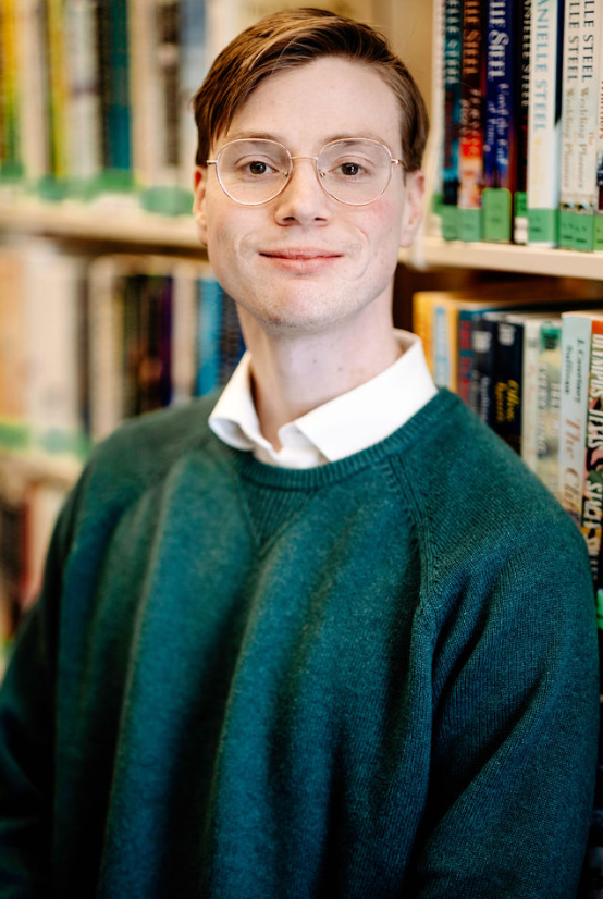 Man in glasses smiles at camera while standing in front of a bookshelf