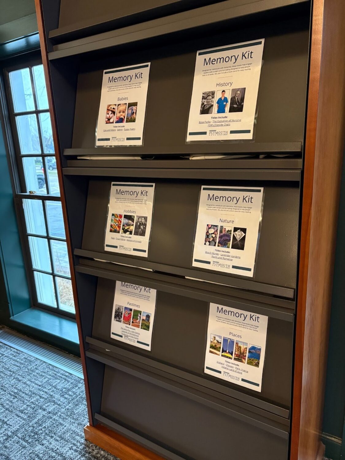 Grey shelves containing memory kits for seniors located near a window on the Main Level at the Plymouth District Library.