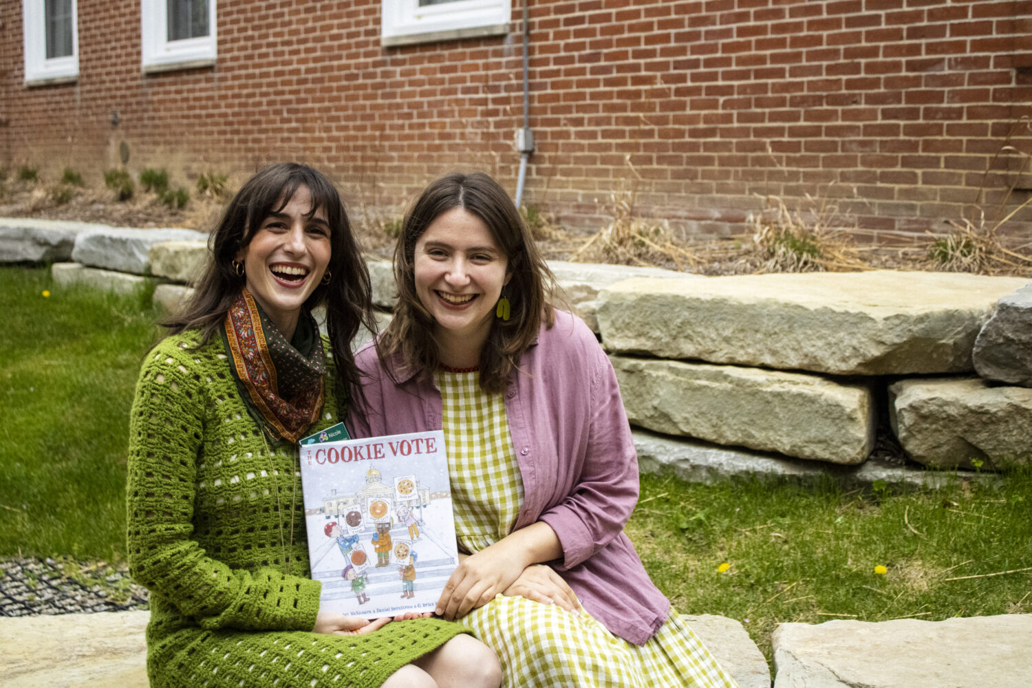 Two librarians sit outside a brick building holding a book, The Cookie Vote.