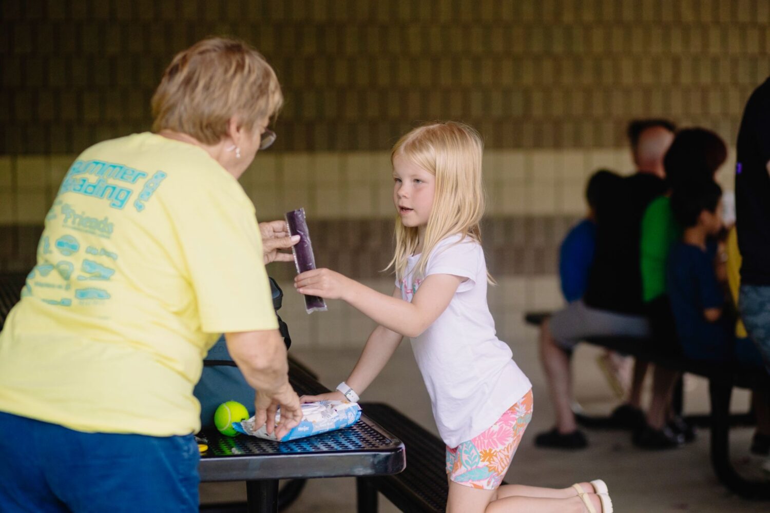 PDL staff member in a yellow shirt hands a purple popsicle to a young girl.