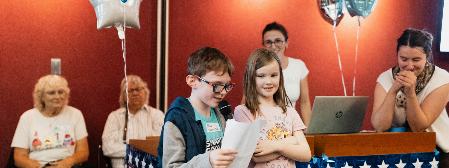Tonquish Toffee bakers, Micah and Quinn Lorentz, a young boy and girl, hold a microphone and piece of paper to give their campaign speech.
