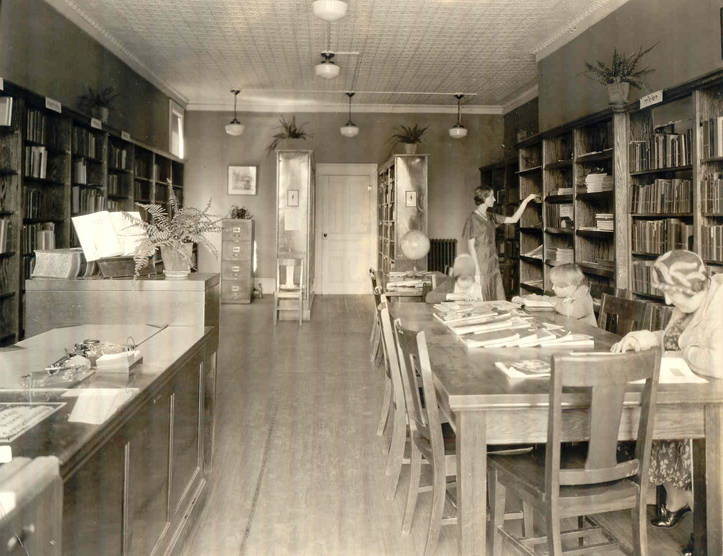 An old-fashioned photo from the 1920s showing a woman shelving books, two children reading at a table with an older woman wearing a shawl.